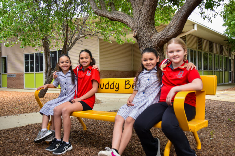 Four primary students sitting together on the school's buddy bench under a tree