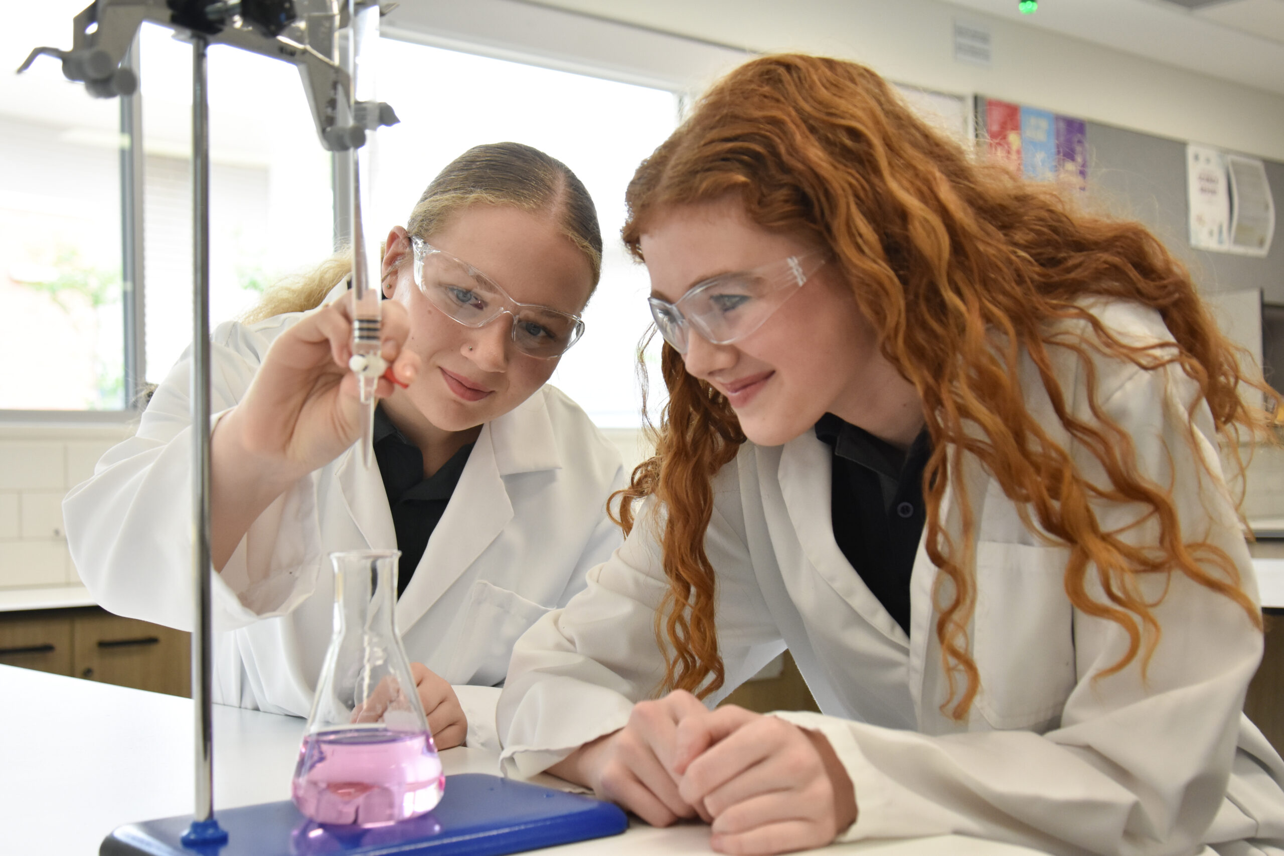 Two students in lab coats and safety glasses running a chemistry experiment