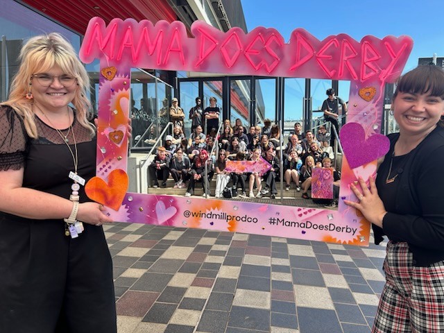 Staff holding a Mama Does Derby photo frame with students in the background